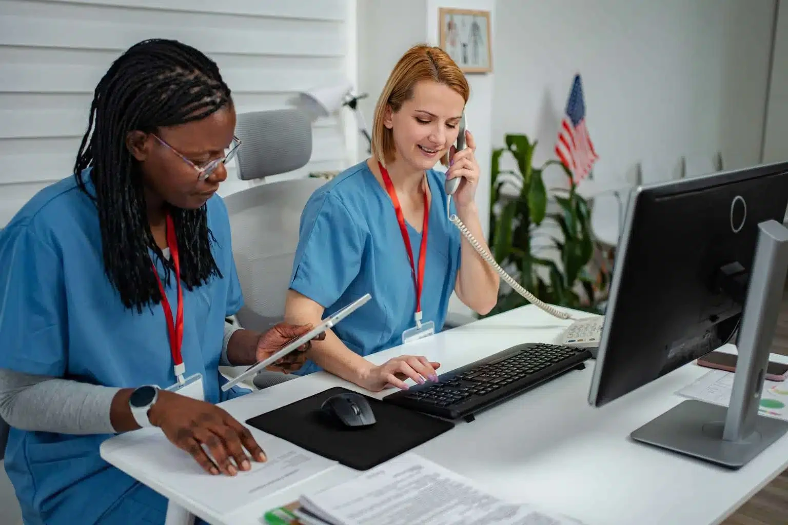 Two medical office staff using a tablet, telephone, and desktop computer at reception