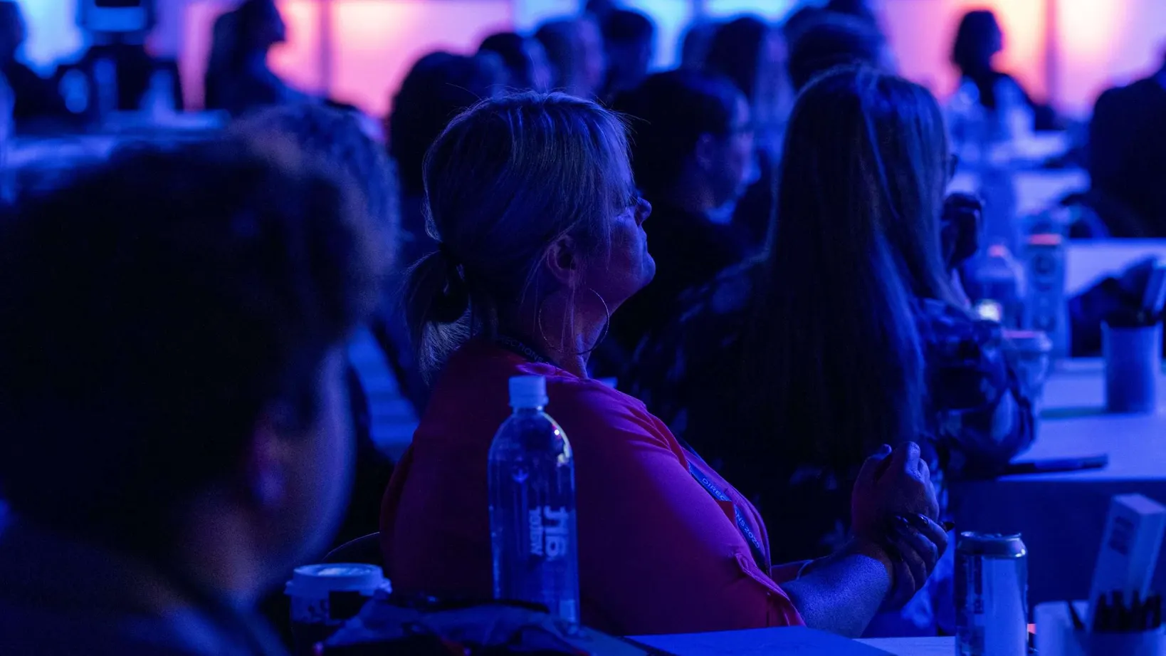Attendees seated at tables in a conference ballroom lit in blue and pink, watching a general session