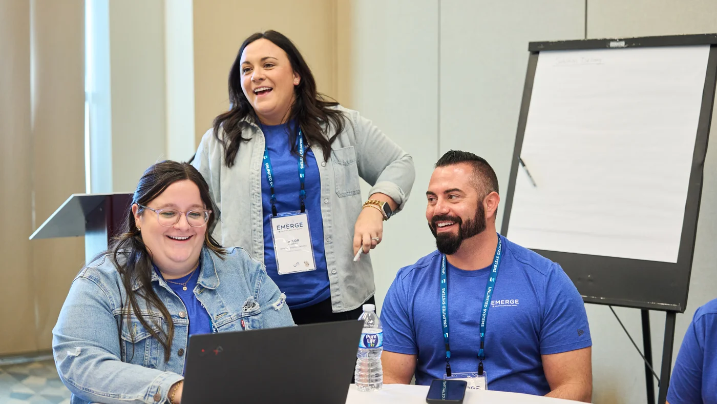 Unlimited Systems team members laughing together while reviewing work on a laptop at the EMERGE Employee Summit