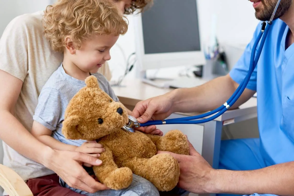 Pediatric clinician listening to a teddy bear's heartbeat for a smiling child on a parent's lap